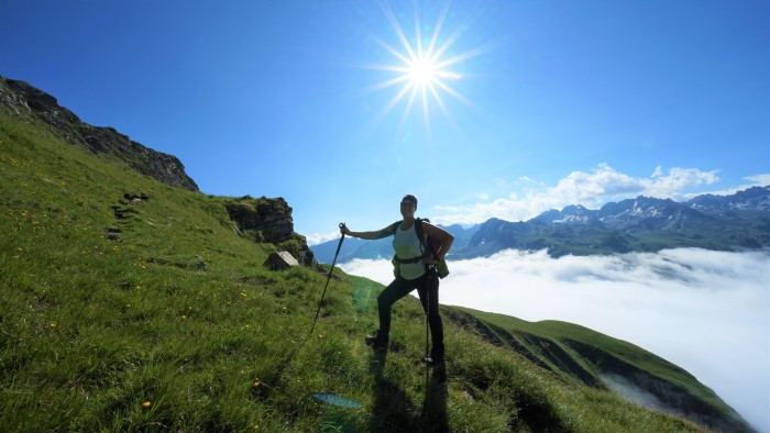 Sabrina Erhard mit Wanderstöcken stehend auf begrüntem Hang, im Hindergrund Berge und dazwischen Sicht auf die Wolkendecke von oben.