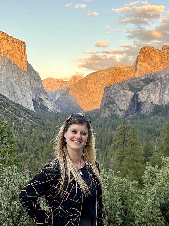 Nicole Müller stehend in der Natur, mit einem imposanten Sonnenuntergang im Gebirge im Hintergrund